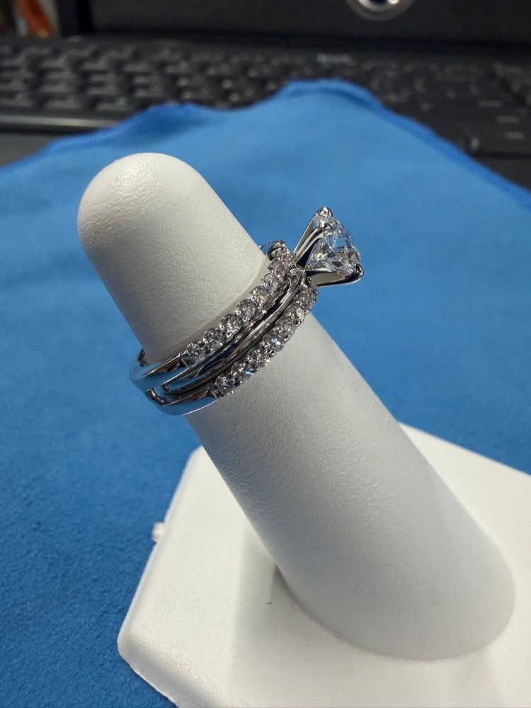 Progress photo of a white metal round solitaire engagement ring with two diamond bands stacked below it, on a ring stand